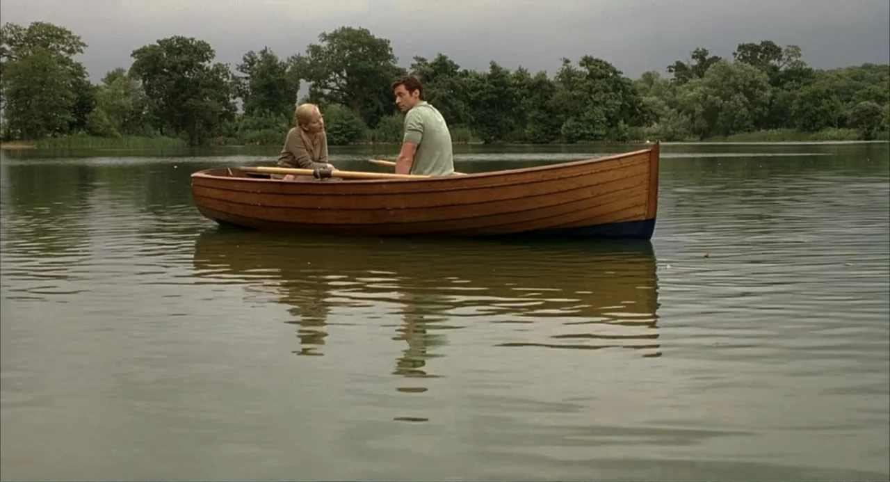 Scoop (2024) movie still: wide — A calm lake is surrounded by a dense green treeline under an overcast sky. A man and a woman sit together in a…