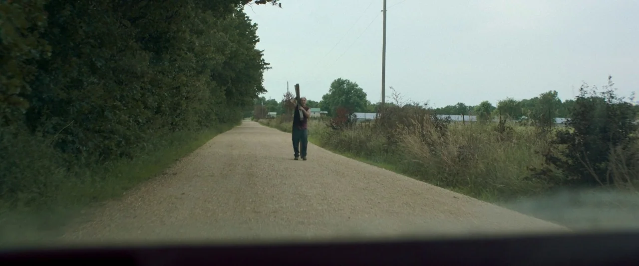 Minari (2020) movie still: wide — A man walks along a rural gravel road bordered by dense trees and fields. He carries a wooden plank on his…