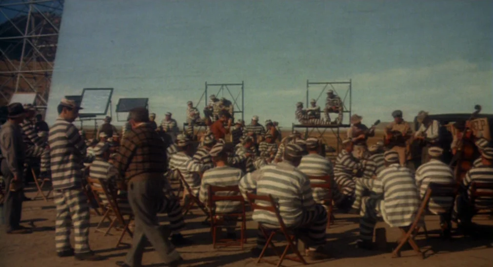 Bound For Glory (1976) movie still: wide — A group of men in striped prison uniforms sit in folding chairs outdoors. Metal scaffolding towers stand…