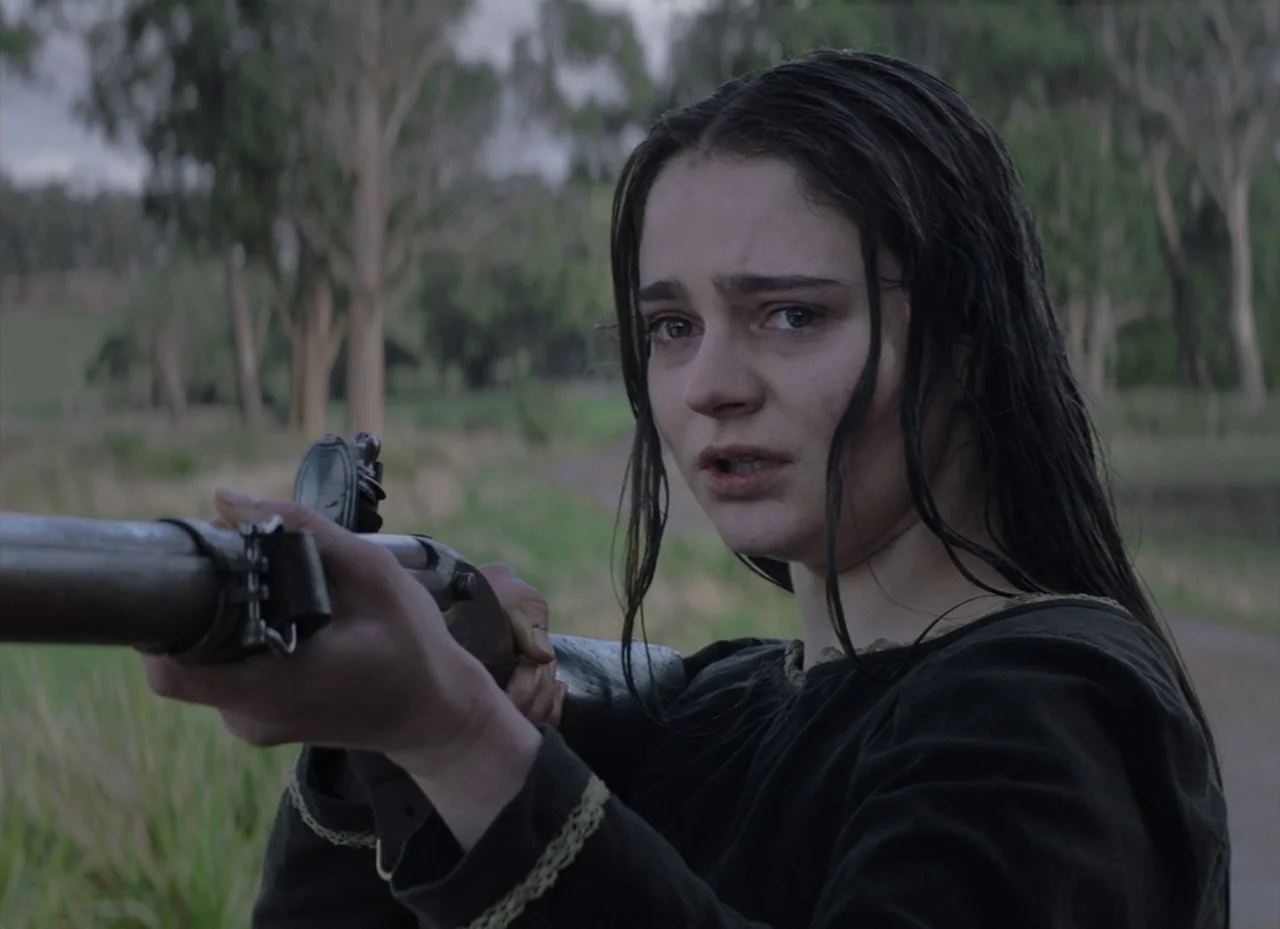 The Nightingale (2018) movie still: medium — A woman stands in a sparse woodland setting with blurred trees in the background. She holds a long…