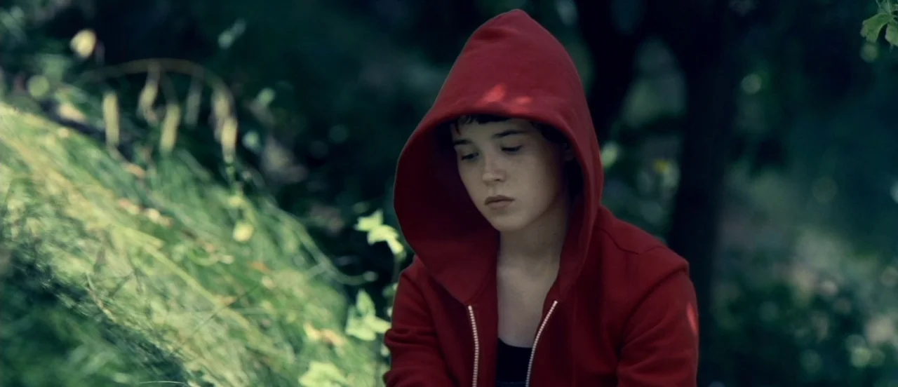 Hard Candy (2005) movie still: medium — A person stands in a dark, shaded patch of a dense green forest. The individual looks down while wearing a…