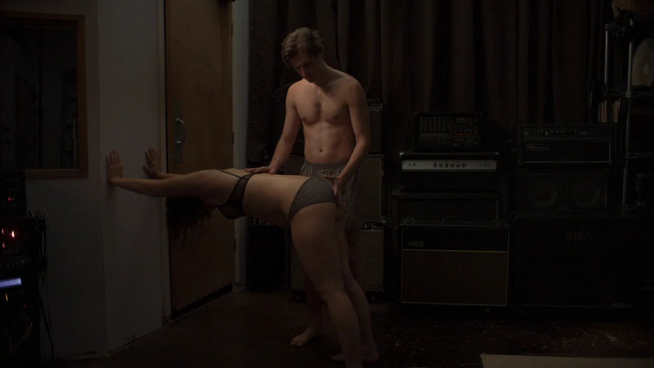 The Feeling That the Time for Doing Something Has Passed (2023) movie still: fullbody — A dimly lit room is filled with stacks of amplifiers and…