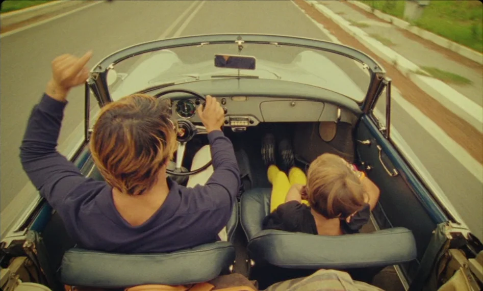 Misunderstood (2014) movie still: medium — A high-angle view from the backseat of a vintage convertible driving down a road. A driver and a young…