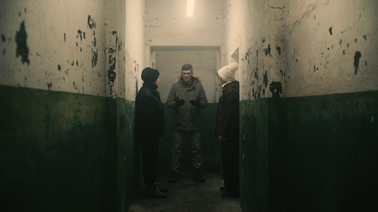 Helll Hole (2024) movie still: fullbody — A narrow, decaying concrete hallway features peeling paint on lower walls with visible grime. Three people…