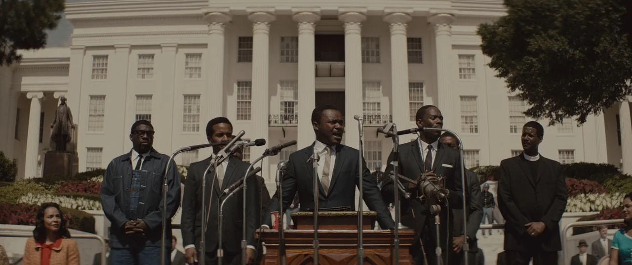 Selma (2014) movie still: wide — A group of men stand before microphones at a podium in front of a white building. The central figure addresses a…