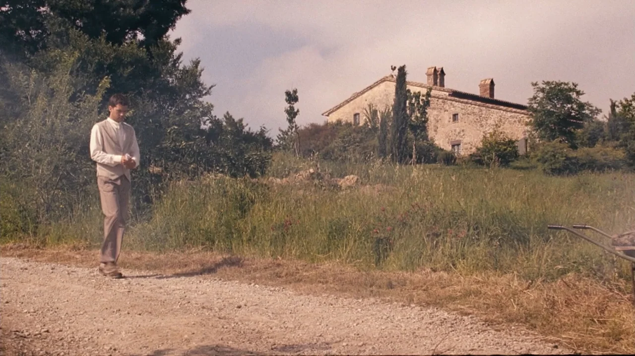 Shadows in the Sun (2005) movie still: fullbody — A rural landscape with a dirt road leading toward a stone farmhouse. A young man walks along the…