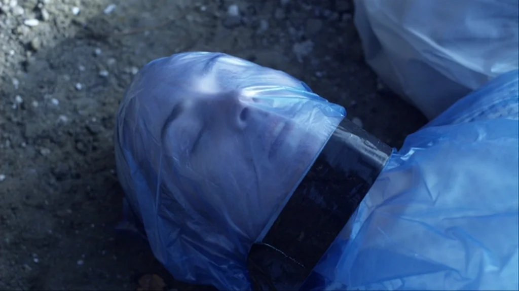 Contagion (2011) movie still: closeup — A person lies on rough dirt ground with a blue plastic sheet wrapped tight. Black tape secures the plastic…