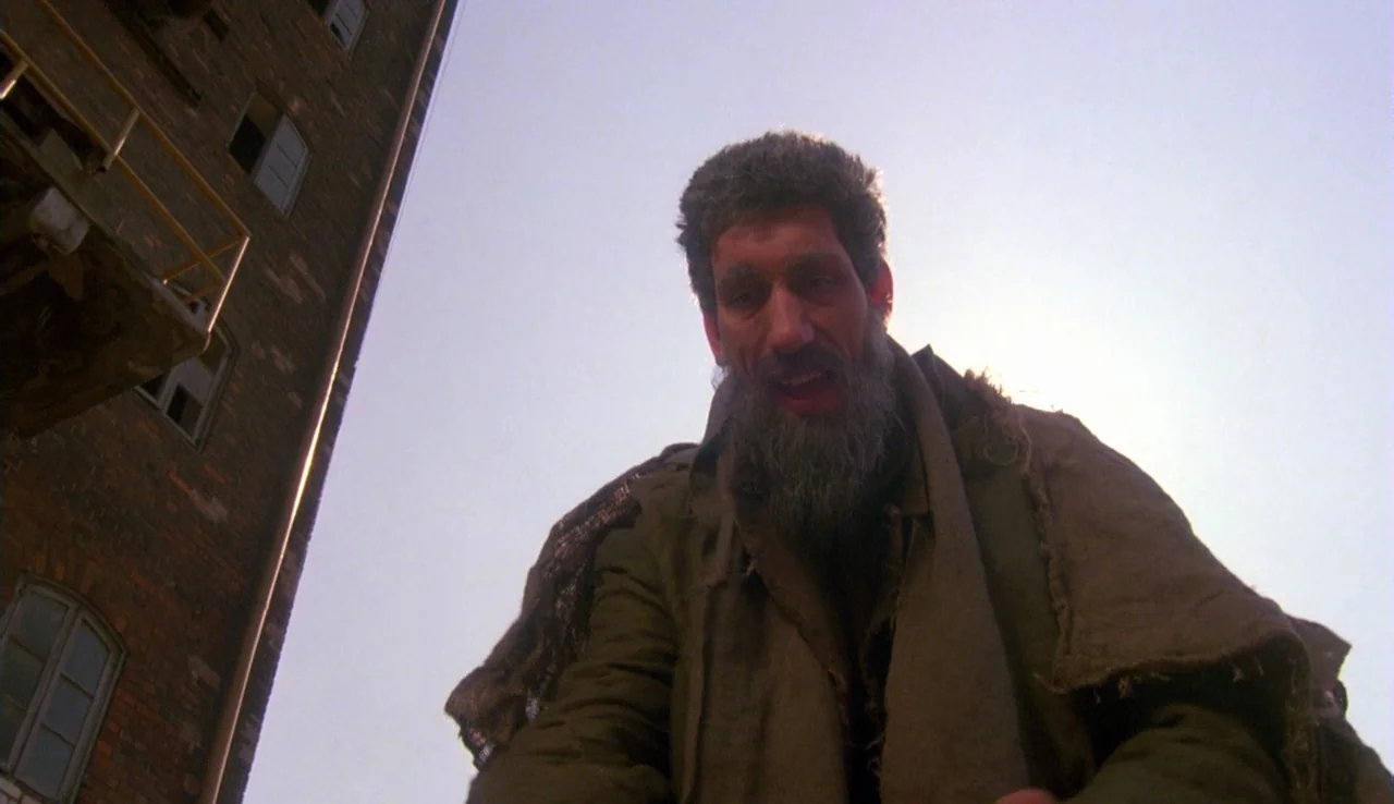 The Rainbow Thief (1990) movie still: medium — A man in a worn cloak leans over an alleyway toward the camera lens. A tall, weathered brick…