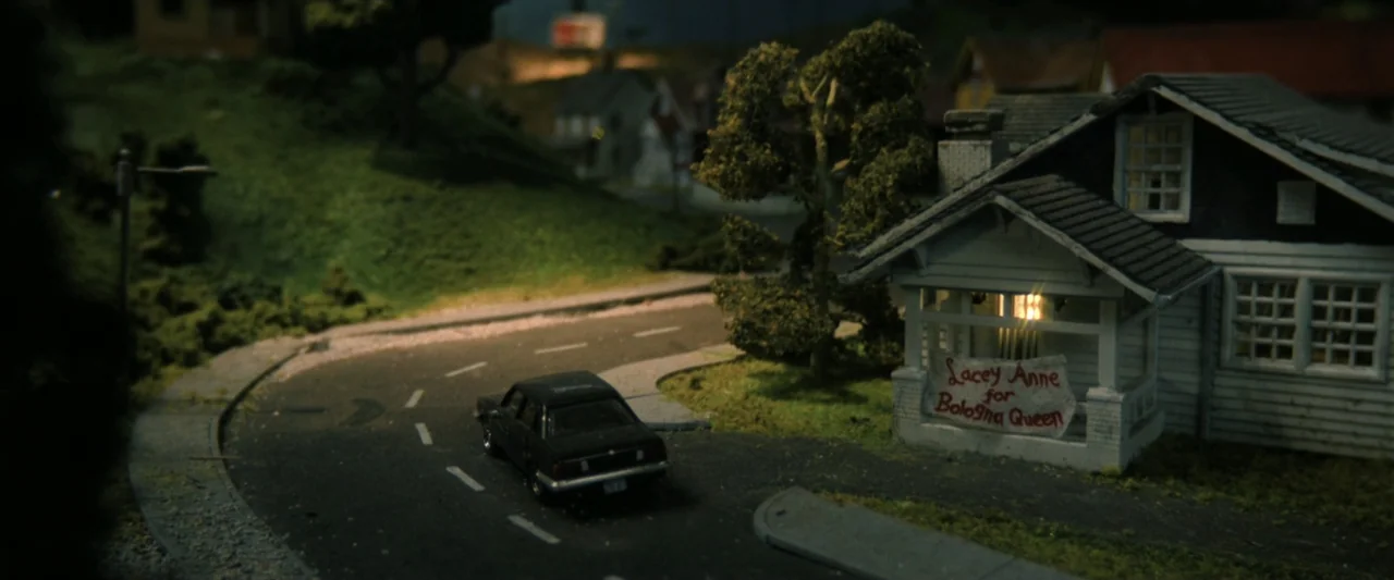 Holland (2025) movie still: establishing — A miniature suburban street scene features a single house and a curved road at night. A black car sits…