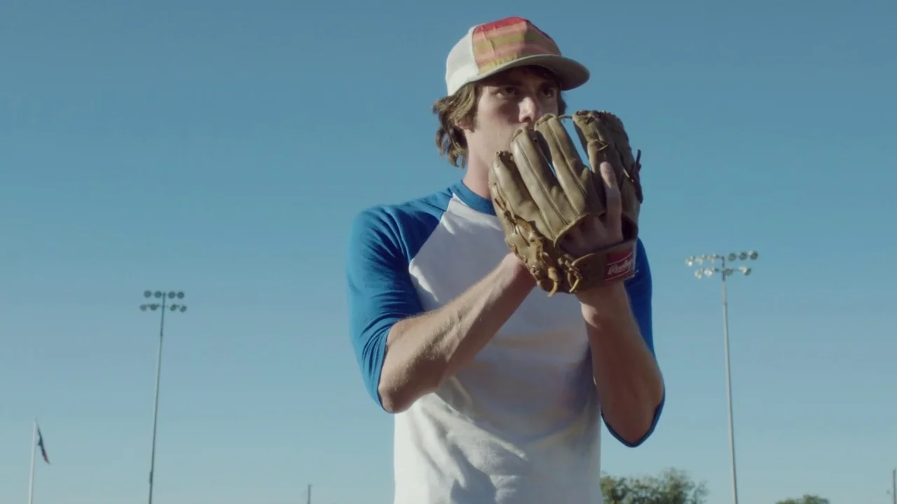 Everybody Wants Some!! (2016) movie still: medium — An outdoor baseball field with tall stadium light poles against a clear blue sky. A young man in…