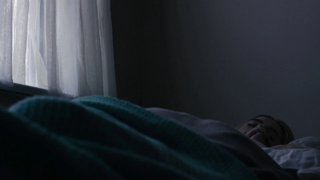 The Blackcoat’s Daughter (2015) movie still: medium — A dark, dimly lit room features a person resting in bed. Light filters through a sheer window…