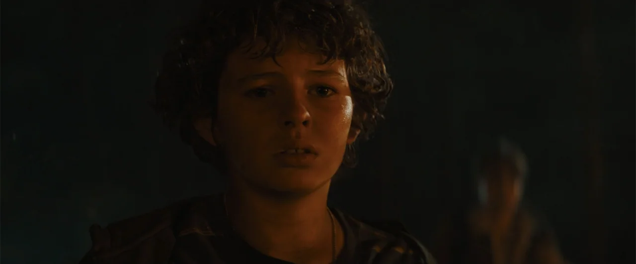 Those Who Wish Me Dead (2021) movie still: closeup — A dark outdoor setting at night with warm orange glow illuminating the subject. A teenage boy…