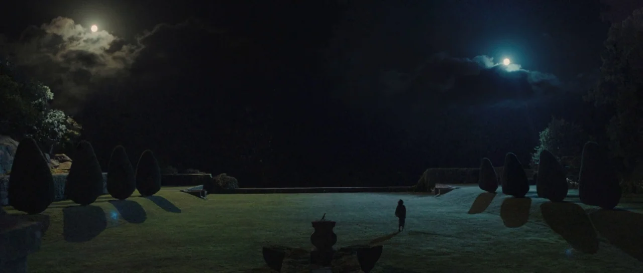 Melancholia (2011) movie still: wide — A vast, open lawn is bordered by symmetrical topiary bushes under a night sky. A lone figure walks across the…