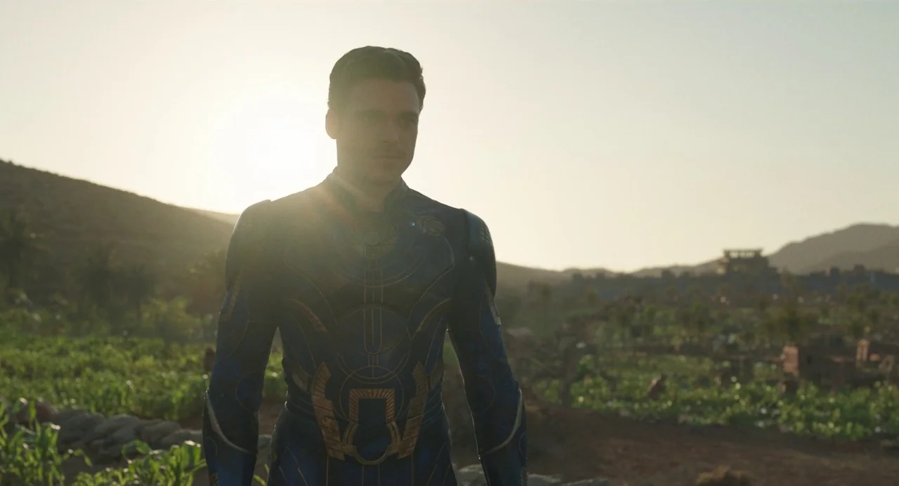 Eternals (2021) movie still: medium — A vast, arid agricultural field stretches toward distant hills under a setting sun. A man wearing ornate blue…