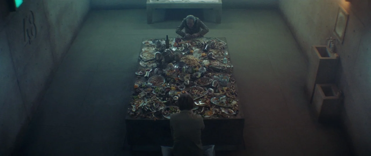 The Platform (2019) movie still: wide — A stark, concrete-walled room features a long central stone platform covered in discarded food. Two men sit…