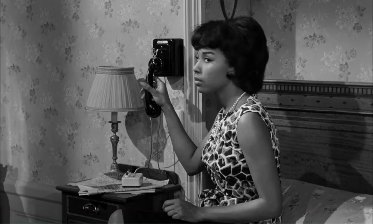 Paris Blues (1961) movie still: medium — A 1960s style bedroom featuring floral wallpaper and a wooden bed frame. A woman sits on the edge of the…