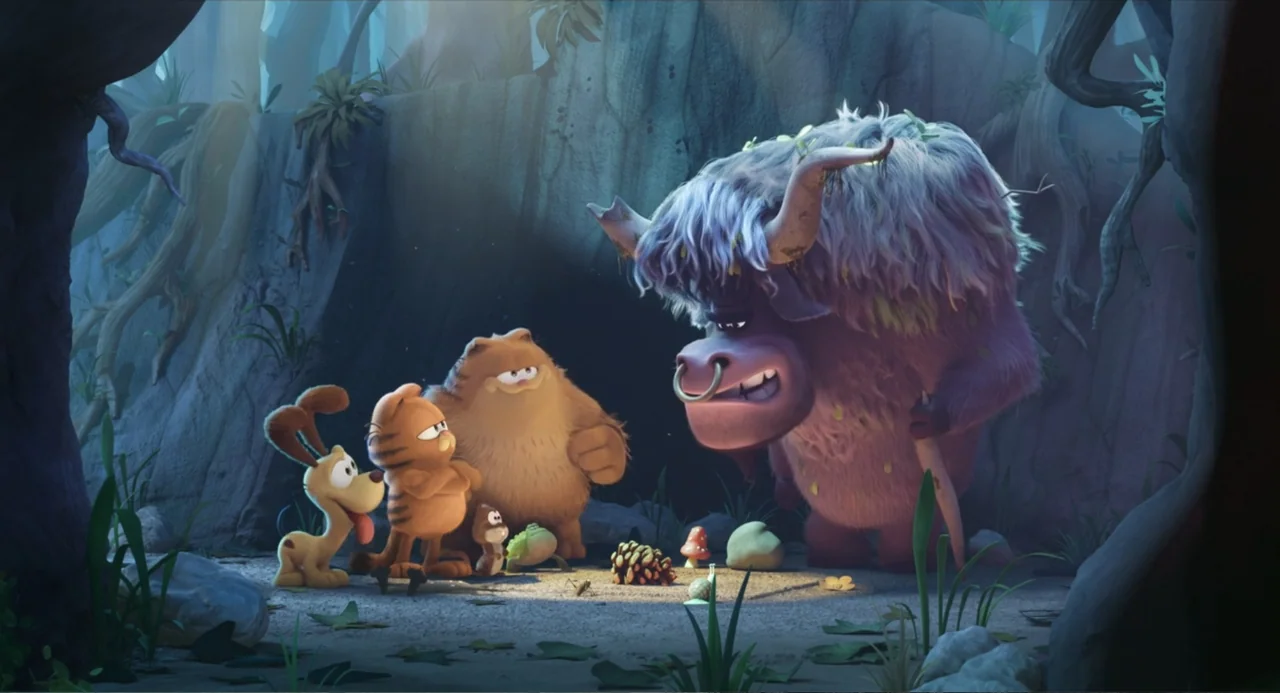 The Garfield Movie (2024) movie still: medium — A rocky cave entrance is surrounded by roots and plants in a forest setting. An orange cat and a…