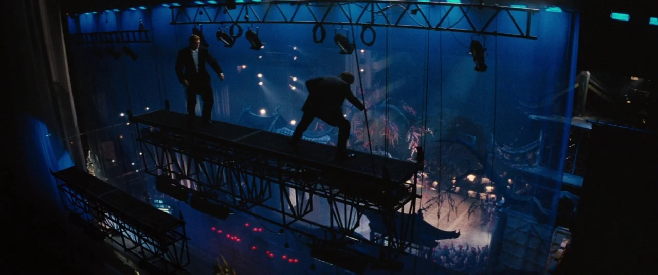 Mission: Impossible – Rogue Nation (2015) movie still: wide — Two men stand on a high metal rigging catwalk suspended above a performance stage. The…