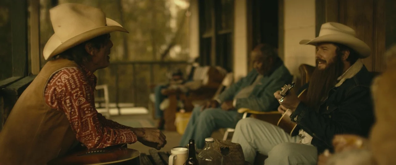 Blaze (2018) movie still: medium — A wooden porch in a rural setting with scattered chairs and beverage bottles. Two men wearing cowboy hats talk…