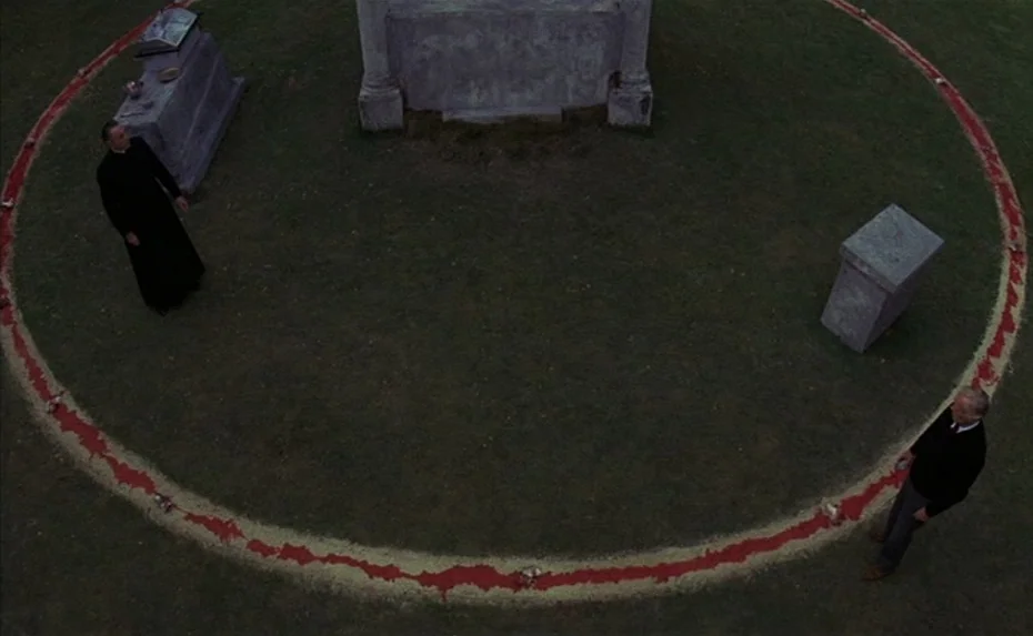 To The Devil a Daughter (1976) movie still: wide — A flat expanse of grass surrounds a large stone monument within a cemetery. Two men are…