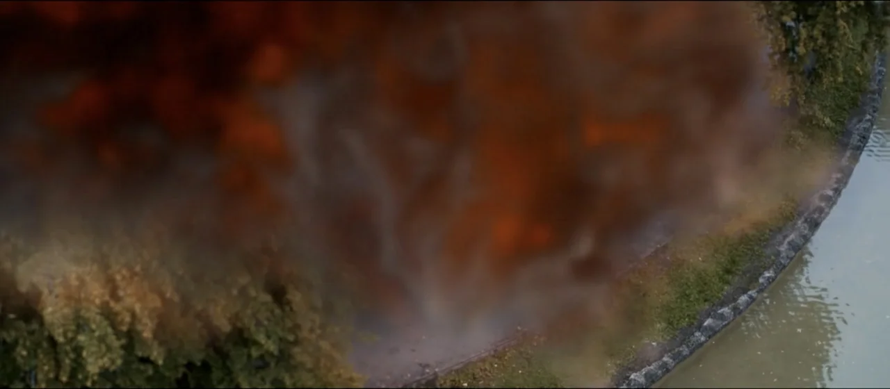 Guns Akimbo (2020) movie still: establishing — An aerial view shows a riverbank bordered by grass and trees with a dark plume of smoke rising from…