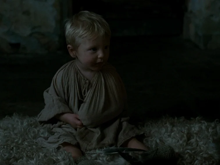 Wuthering Heights movie still: medium — A dark, dimly lit interior room features stone walls and a sheepskin floor covering. A small blonde child…