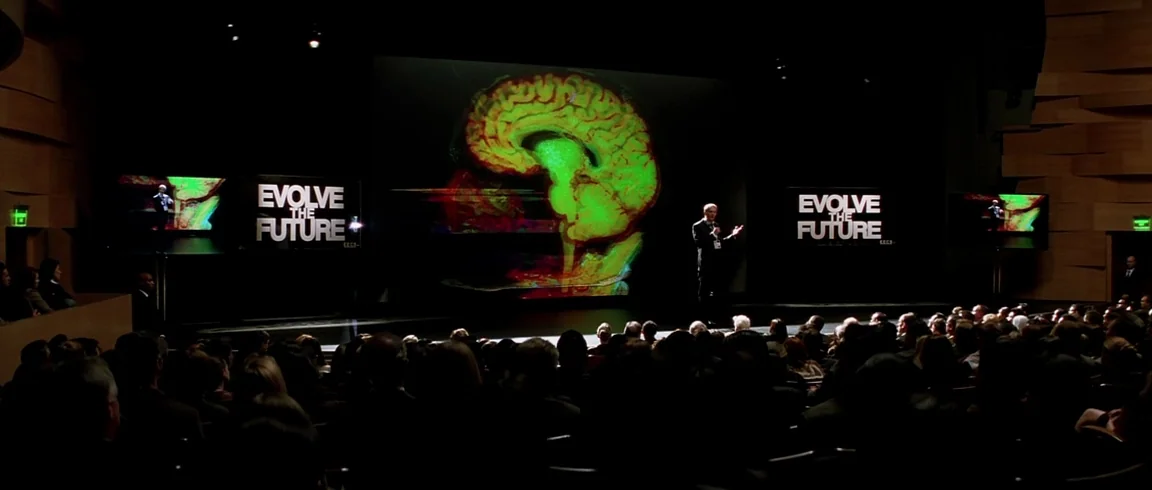 Transcendence (2014) movie still: wide — A dark, modern auditorium features a man lecturing in front of a giant digital display. The screen depicts…