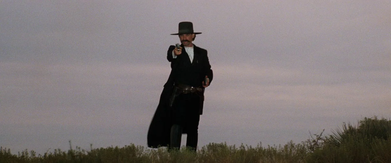 Tombstone (1993) movie still: fullbody — An open rural landscape under a flat, overcast sky during twilight. A mustached man in period-accurate…