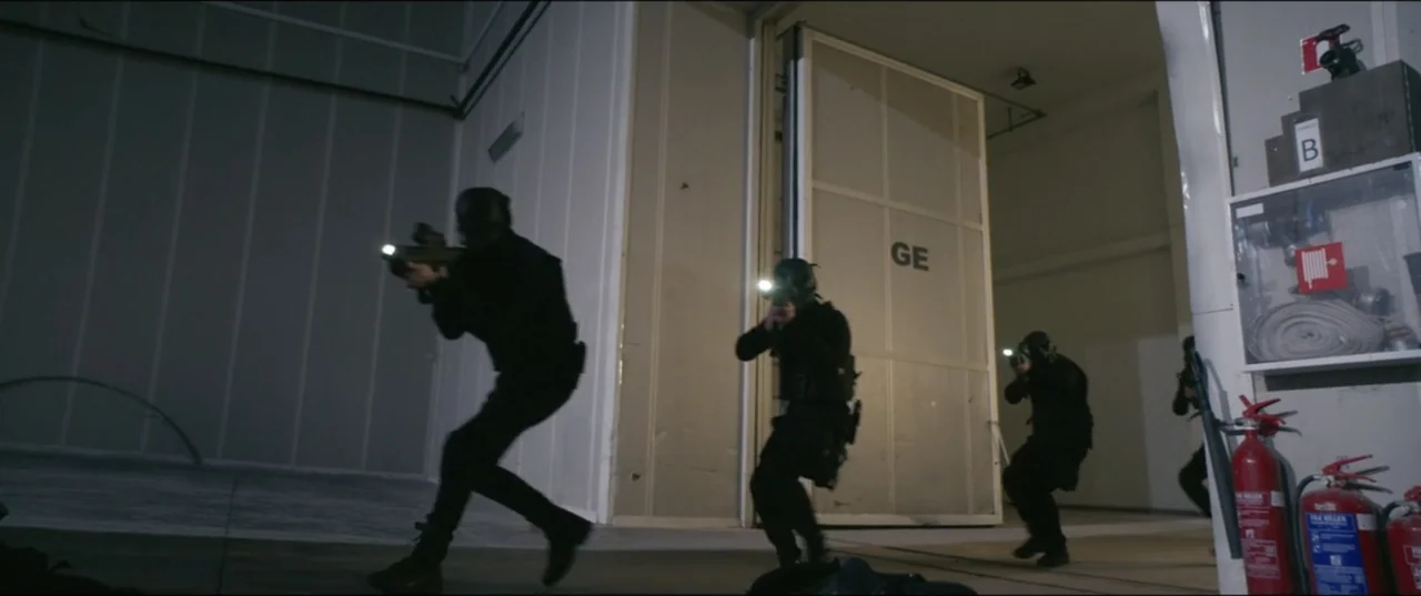 Utopia (2013) movie still: fullbody — An industrial hall with white paneled walls and large open sliding metal doors. Four soldiers in black…