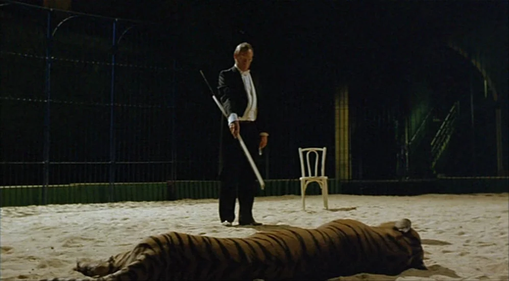 Roselyne and the Lions (1989) movie still: wide — A dimly lit sand-covered ring is surrounded by tall metal fencing. A man in formal attire stands…