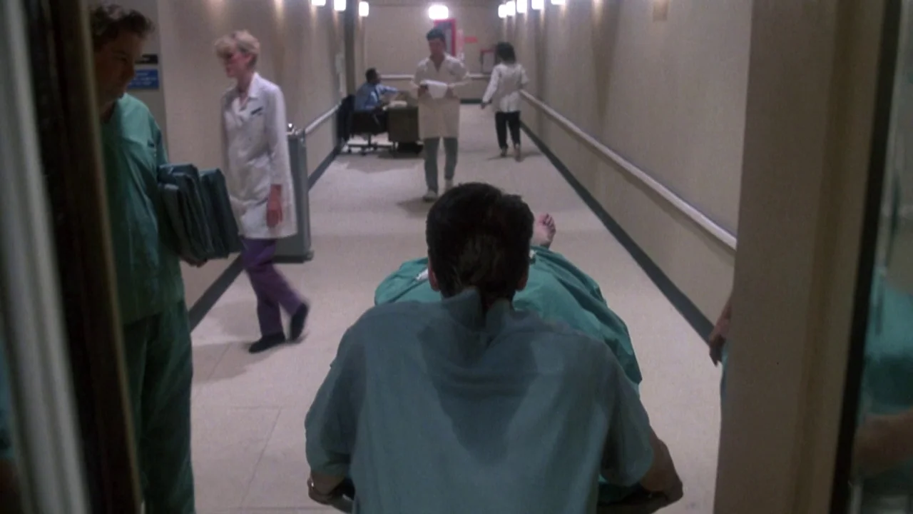 Re-Animator (1985) movie still: medium — A long, brightly lit hospital corridor stretches into the distance with beige walls. A man in green scrubs…