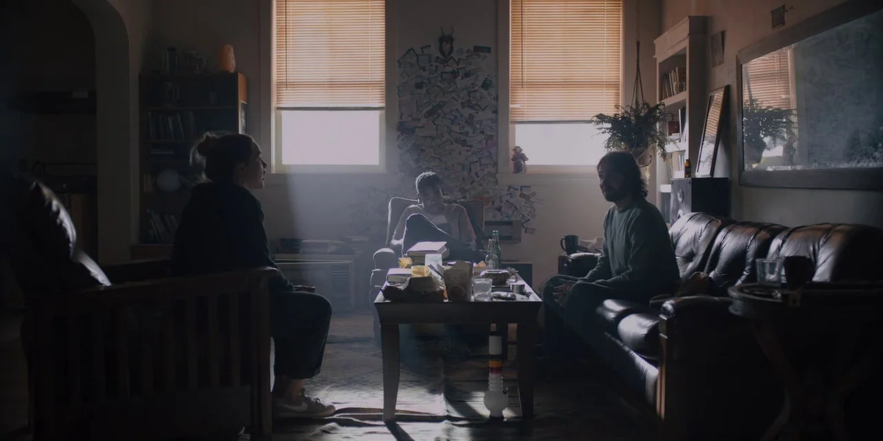Midsommar (2019) movie still: wide — A cluttered apartment with wooden furniture, blinds over two windows, and miscellaneous personal items. Three…