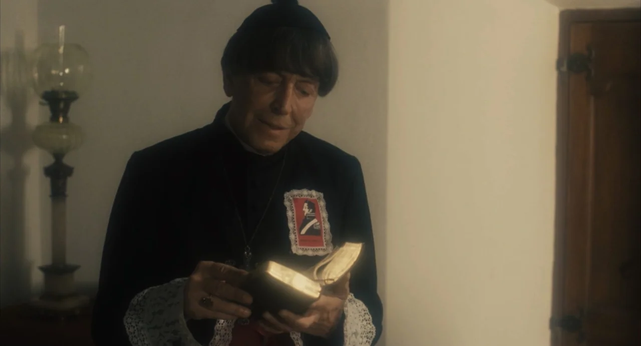 Camila (1984) movie still: medium — An interior space with a beige wall and a vintage lamp in the background. An elderly man in clerical attire…