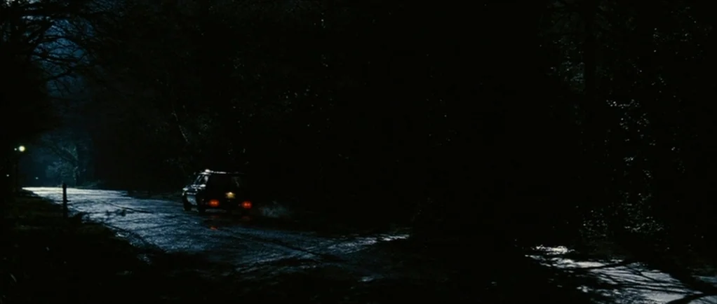 The Walker (2007) movie still: wide — A dark, narrow country road is lined with dense trees on both sides at night. A car with visible red…