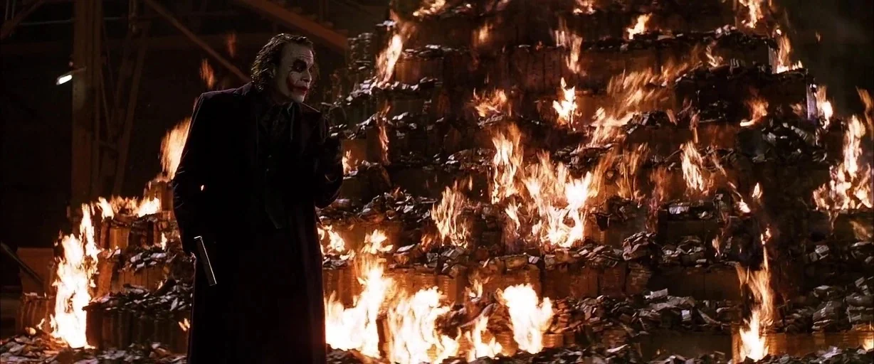 The Dark Knight (2008) movie still: medium — A man stands before a massive, burning pyramid made of stacks of cash. The environment is dark and…