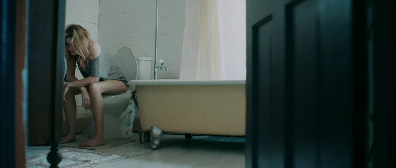 Starfish (2018) movie still: medium — A small bathroom with a clawfoot bathtub and simple white walls. A young woman sits on the toilet facing…