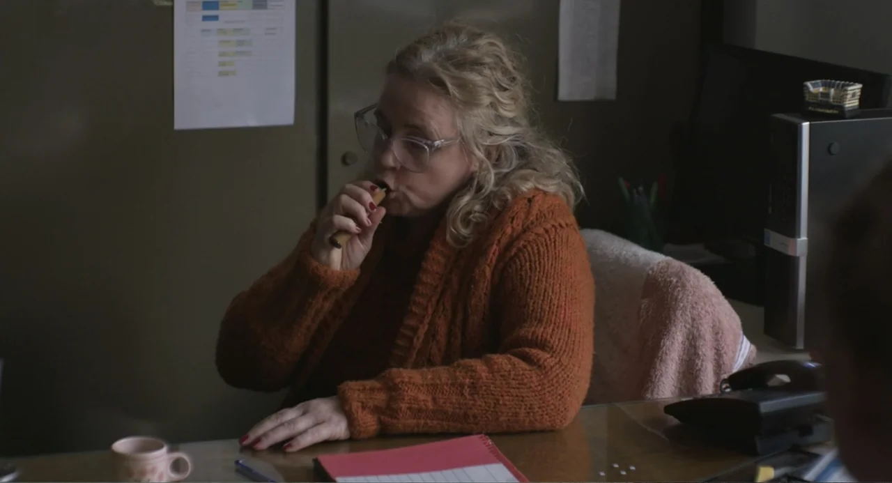 An Odd Turn (2024) movie still: medium — An office interior features a wooden desk with a paper notebook and computer equipment. A middle-aged woman…