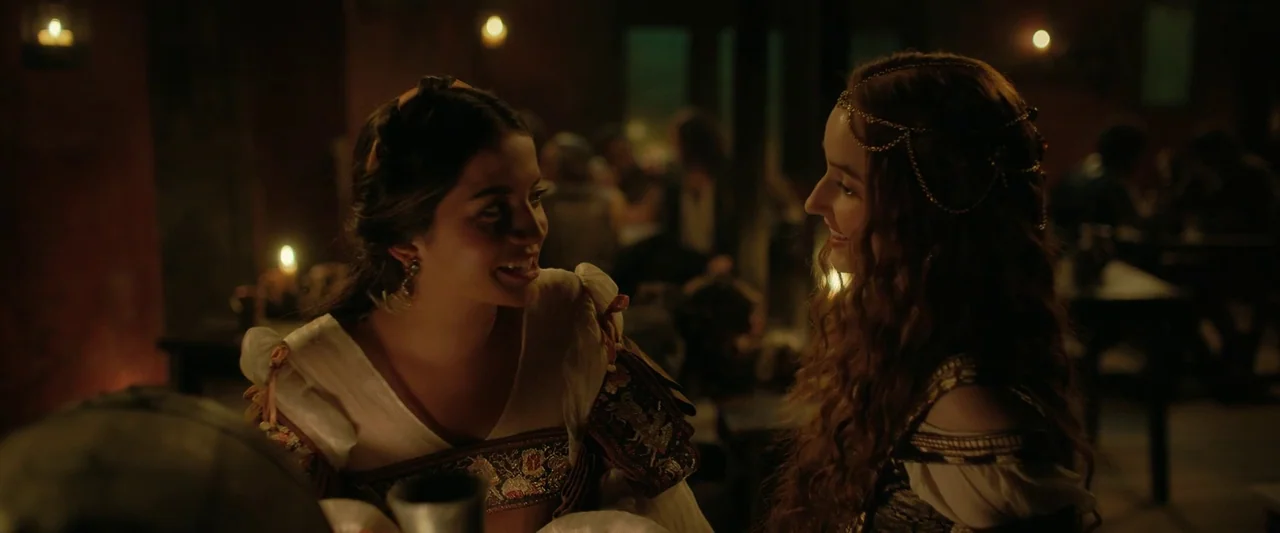 Rosaline (2022) movie still: medium — A dimly lit medieval-style tavern interior with warm candles providing the primary light source. Two women…