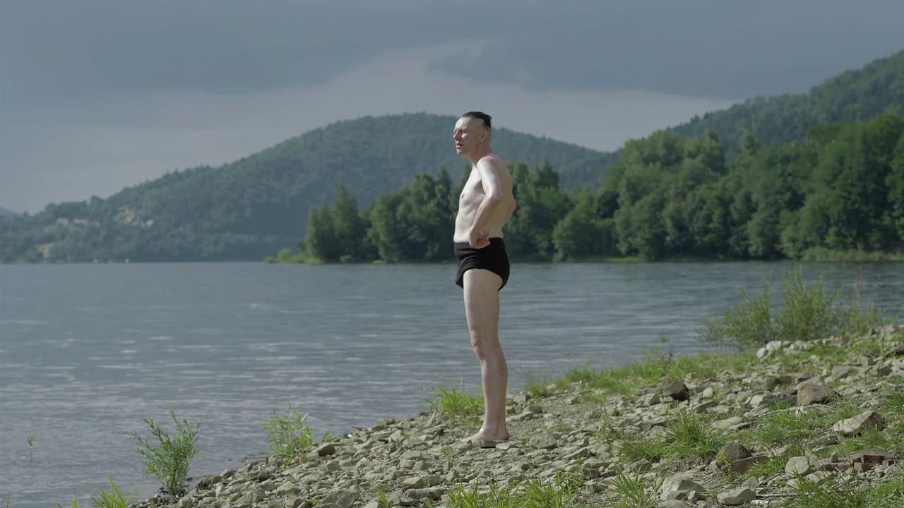 The Zone Of Interest (2023) movie still: fullbody — A rocky, grassy bank borders a wide lake under an overcast sky. A man in black swim trunks…