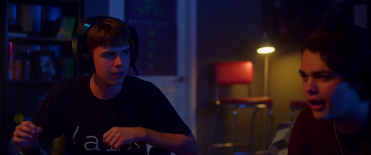 Blindfire (2020) movie still: medium — A dimly lit room contains two young men sitting in front of computer screens. A red barstool stands in the…