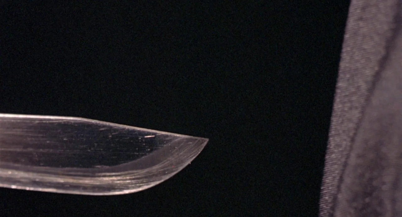 Ms. 45 (1981) movie still: closeup — A close-up view shows the metallic, reflective edge of a blade against darkness. A textured black fabric…