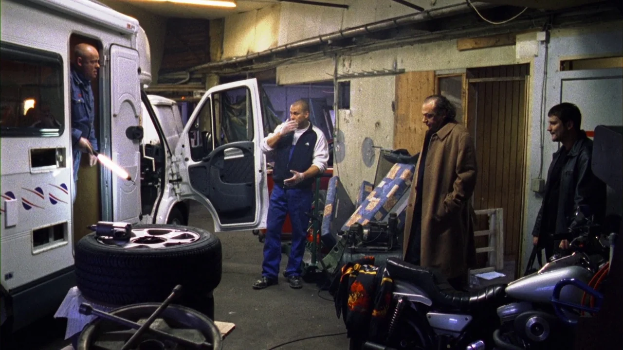 Pusher III (2004) movie still: wide — A cluttered mechanic workshop features a white camper van with an open side door. Four men stand in the space…