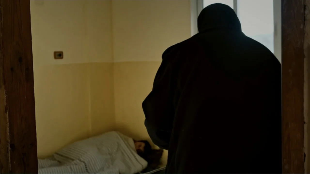 No Other Land (2024) movie still: medium — A spartan room features beige walls and a person sleeping in a bed. A hooded figure stands in the…