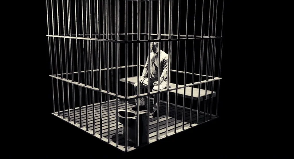 Sin City (2005) movie still: fullbody — A man sits alone inside a metal cage structure set against a black background. The cage has a wooden bench…