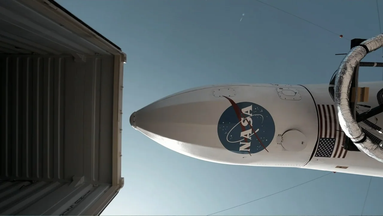 Wild Wild Space (2024) movie still: wide — A tall white rocket displays a large NASA logo under a clear blue sky. The vehicle is positioned next to…