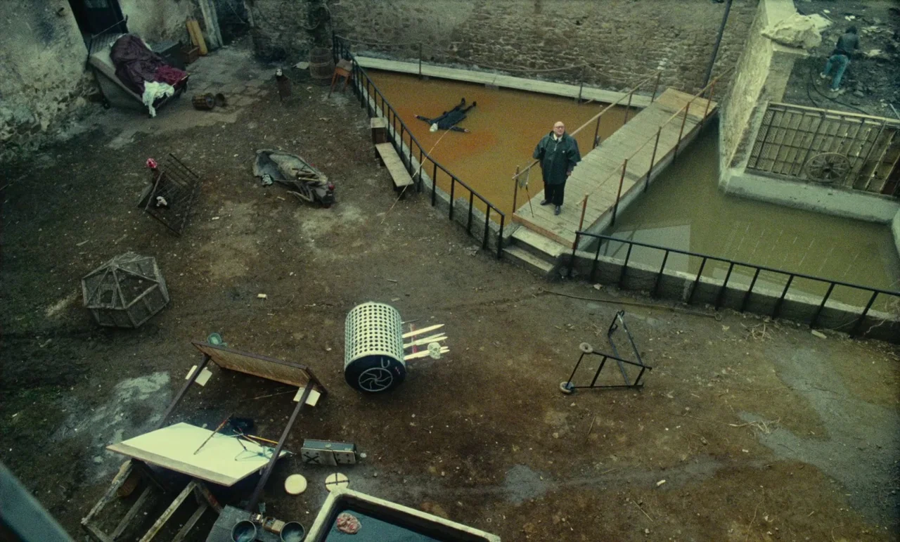 Litan (1982) movie still: wide — An open muddy courtyard features a triangular pit of brownish liquid with a walkway. An older man stands on the…