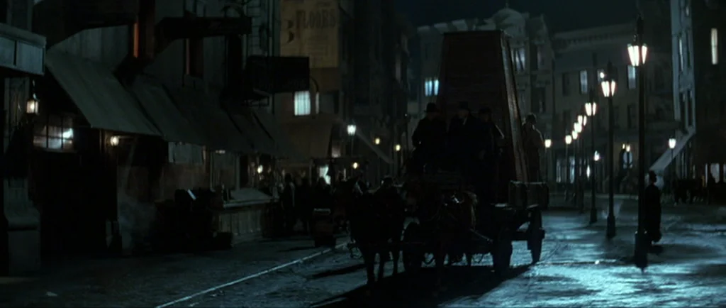 The Prestige (2006) movie still: wide — A dark, wet urban thoroughfare is lined with old buildings and rows of streetlamps. A horse-drawn wagon…