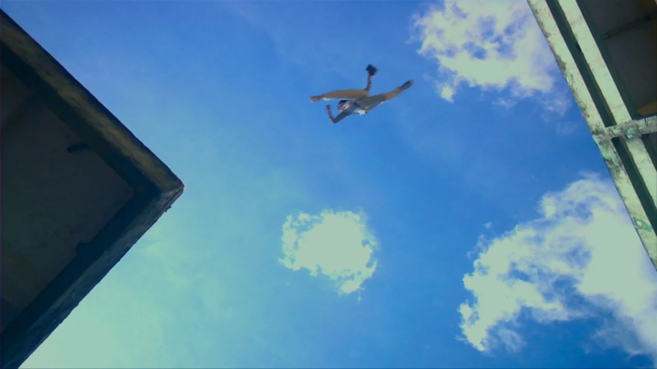 Merantau (2009) movie still: wide — Two concrete building corners frame a clear blue sky with sparse white clouds. A person is suspended in mid-air…
