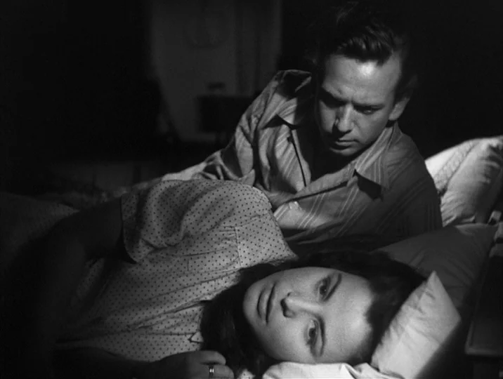 To Joy (1950) movie still: medium — A dark, dimly lit room contains two people lying close together in a bed. The man rests on his side behind the…