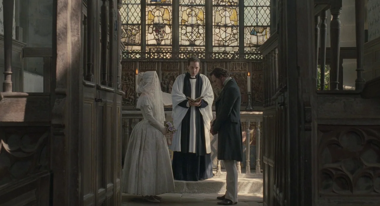 Jane Eyre (2011) movie still: fullbody — A stone church interior features carved wooden pews and a stained glass altar backdrop. A priest officiates…